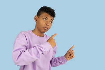 Dark skinned Latino teenager wearing a purple sweatshirt pointing with his index fingers at a copyspace on a light blue background, young man indicating an advertising space