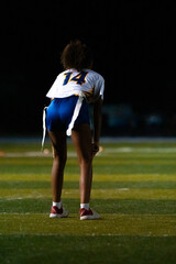 A female flag football player in ready stance under stadium lights. Number 14 on defense, focused and alert before the snap during a competitive night game.