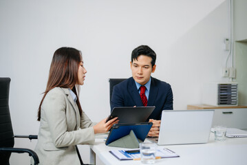 Fototapeta premium Two Asian business professionals in formal suits discussing a project at modern office desk with laptop and documents.