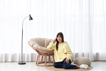 Young woman talking by mobile phone on rug near armchair at home