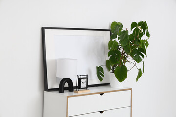 Monstera houseplant, lamp and clock on chest of drawers near white wall in room, closeup
