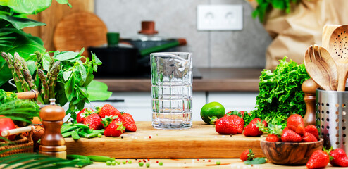 Green basil, lime and fresh strawberries for making summer non alcoholic cocktail drink in glass on wooden table, preparing a healthy vegan meal in a cozy kitchen.