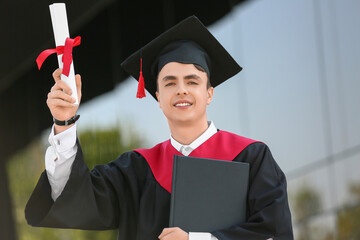 Male graduating student with diploma and book near university