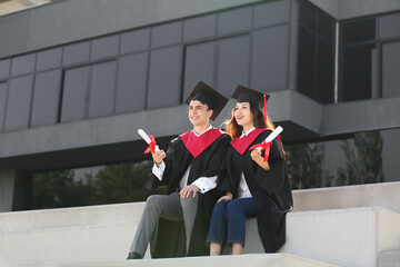 Couple of happy graduating students with diplomas sitting on steps outdoors