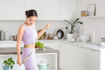 Sporty young Asian woman measuring her waist in kitchen. Diet concept