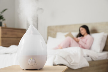 Air humidifier on table against woman reading book in bedroom, closeup