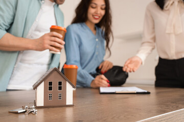 House model and keys on table against real estate agent with couple signing agreement in kitchen,...