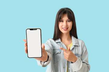 Smiling young woman pointing at blank mobile phone on blue background