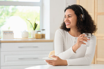 Young African-American woman with headphones and glass of water using mobile phone at table in...