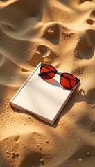 A closed white book and red-tinted sunglasses rest on rippled golden sand under warm sunlight at a beach.