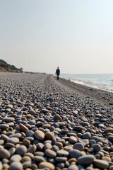 A solitary person walks along a pebble-covered beach beside calm sea waters under a clear sky.