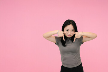 short asian woman pointing down with dissapointed expression in pink studio background