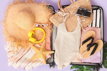 Open suitcase with clothes, swimsuit, beach accessories and photo camera on lilac background, closeup