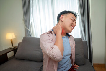 Indonesian southeast asian man suffering from neck pain while sitting on the living room couch at...