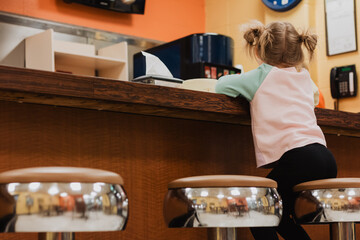 Young child leaning over wooden café counter in pastel sweatshirt with space buns hairstyle at a retro-style diner interior under warm ambient lighting and solitude