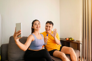 Indonesian southeast asian couple drinking water while taking a selfie or video call with their cellphone after finishing a home body workout exercise session. Fitness and active lifestyles concept