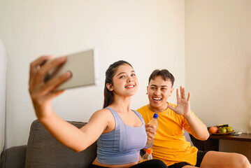 Indonesian southeast asian couple drinking water while taking a selfie or video call with their cellphone after finishing a home body workout exercise session. Fitness and active lifestyles concept