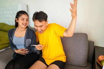 Indonesian southeast asian couple sitting on a sofa at home while looking results of a pregnancy test. Healthcare and family planning concept