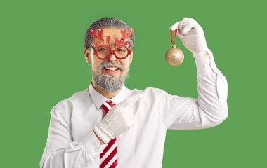 Handsome man dressed in festive clothing, pretending being Santa Claus, pointing at Christmas tree decoration, isolated over green background. New Year is coming soon, think about decor