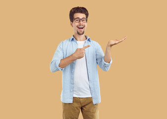 Laughing happy bearded man in glasses pointing index fingerand hand on copy space on beige background. Banner for advertisement, marketing, sales, offer, discount. Sincere human emotions concept.