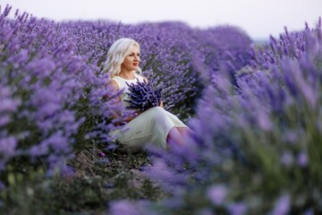 Blonde woman poses in lavender field at sunset. Happy woman in white dress holds lavender bouquet. Aromatherapy concept, lavender oil, photo session in lavender