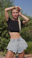 Blonde tanned Caucasian woman with navel piercing wears denim shorts and black cropped top in a park