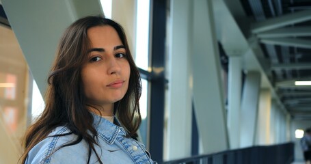 Young woman with long brown hair, wearing a denim shirt, sips coffee from a disposable cup in a modern building with large windows and metal framework