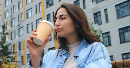 Stylish young woman sits on a bench, savoring a hot beverage from a disposable cup, taking a break amidst the backdrop of a modern building