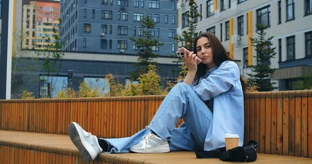 Young woman, wearing casual attire, sits on a wooden bench, savoring a takeaway coffee while engaging with her smartphone against a backdrop of modern buildings