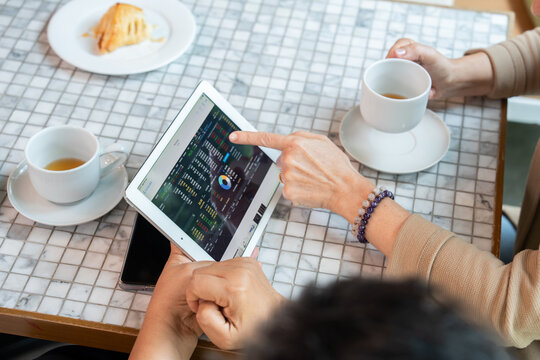 Hands of mature women analyzing colorful financial chart and data on tablet at tiled cafe table. One pointing at screen while the other holds coffee. Plates with pastry and white coffee cups present. - Powered by Adobe