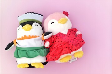 Two adorable penguin plush toys dressed in colorful costumes—one in a green and white outfit and the other in a red polka-dotted dress—lie against a pastel pink background.