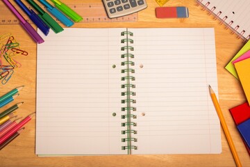 Overhead view is showing open spiral-bound notebook on wooden desk with pens, markers, calculator