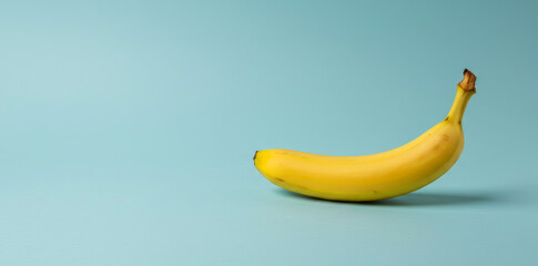 A single yellow banana lies on a light blue surface in a studio.