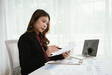 A young Asian businesswoman in a black blazer and red shirt calculates numbers on a white...