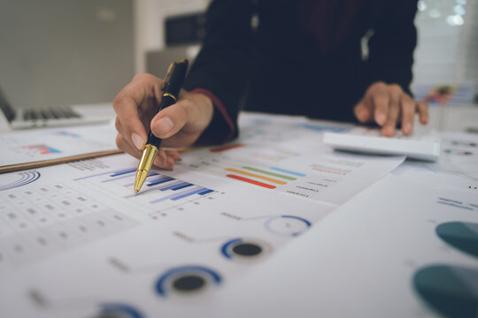 A businesswoman analyzes financial documents with charts and graphs at a desk, highlighting data trends with a pen in a bright, professional office setting.