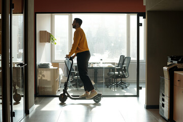 Young Black man riding electric scooter through modern coworking office space, wearing yellow sweater and glasses, passing by glass-walled room © pressmaster
