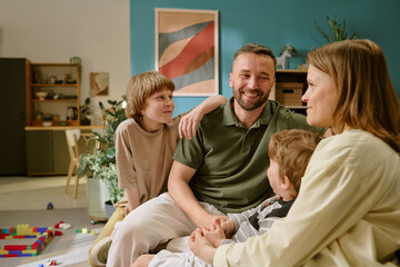 Caucasian middle aged man sitting with Caucasian woman, young boy and child boy, smiling and talking together in home living room, colorful toys scattered on floor, work from home context