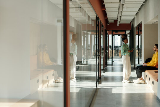 Young Black woman standing and talking to biracial man in modern coworking space with glass walls, both wearing casual clothing