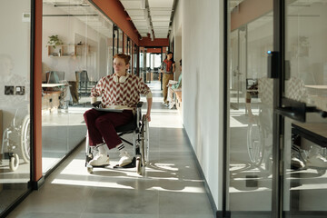 Young Caucasian man with short red hair using wheelchair moving through modern office hallway, two young adults visible in background
