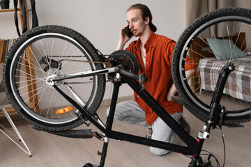 young man talking on smartphone while fixing a bike bicycle at home indoors