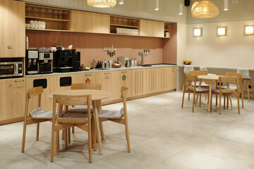 Modern coworking kitchen interior featuring wooden tables and chairs, built-in cabinets, coffee machines, microwave, and utensils on wall, no people visible in workspace