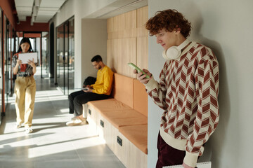 Caucasian man wearing headphones leaning against wall using smartphone while Asian woman and Black...