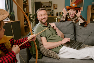 Caucasian middle aged man working on laptop from home, sitting on sofa smiling while two children dressed as cowboys playing around him, one aiming toy bow and arrow, other making playful face