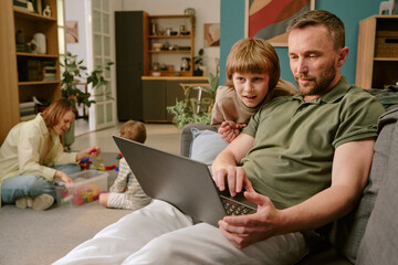 Caucasian middle aged man using laptop on sofa with Caucasian boy leaning over shoulder, woman and toddler playing with toys on floor in background, family working from home together