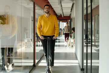 Young man riding electric scooter through modern coworking office hallway, wearing yellow sweater and glasses, people collaborating in background