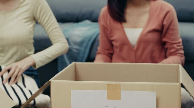 Two asian women organizing folded clothes into a cardboard donation box at home. The image captures a meaningful gesture of giving, charity, and community support.