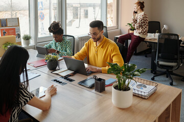 Man in yellow sweater typing on laptop at table with young adult Asian woman and young adult Black...
