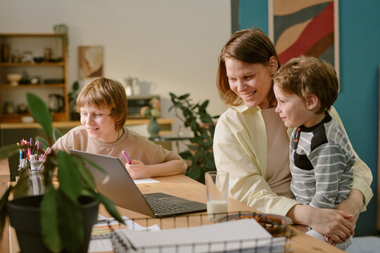 Caucasian middle aged woman working from home on laptop while holding young Caucasian boy, Caucasian child sitting at desk drawing with colored pencils, home office environment - Powered by Adobe