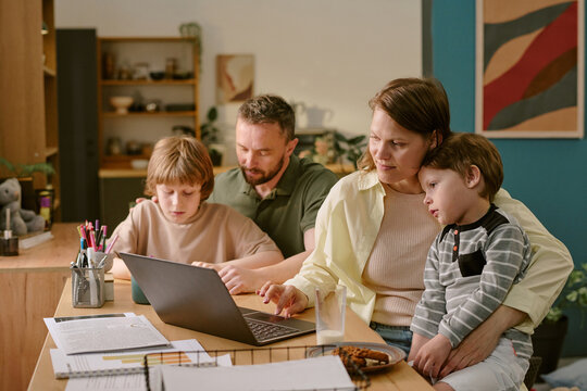 Caucasian middle aged woman working on laptop with young Caucasian boy on lap, Caucasian middle aged man assisting Caucasian child with homework, family multitasking during work from home - Powered by Adobe