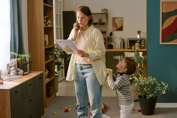 Caucasian middle aged woman talking on smartphone while holding documents, young Caucasian boy tugging at her shirt in home setting, toys scattered on floor, remote work concept